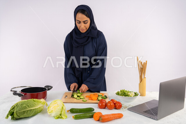 إعداد السلطات والاطباق الصحية، تعلم مهارة الطهي عبر الحاسب المحمول، بورتريه لسيدة عربية خليجية اماراتية محجبة  ترتدي عباءة تقوم بتحضير وصفة طبق صحي مفيد من الانترنت، ربة منزل سعودية تقوم بتقطيع الخضراوات الطازجة، خلفية بيضاء
