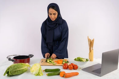 Preparing salads and healthy dishes, learning cooking skills via laptop, portrait of a veiled Emirati Gulf Arab woman wearing an abaya preparing a healthy dish recipe from the internet, a Saudi housewife cutting fresh vegetables, white background