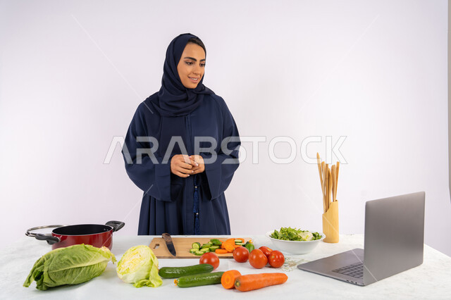 تطبيق وتعلم فنون الطهي من خلال المواقع الإلكترونية، توعية المجتمع بإعداد الطعام الصحي، بورتريه لامرأة عربية خليجية اماراتية محجبة ترتدي العباءة تقوم بتصوير وعرض الطبخ عبر اللابتوب، ربة منزل سعودية تشرح المكونات والكميات المطلوبة عبر بث مباشر، خلفية بيضاء