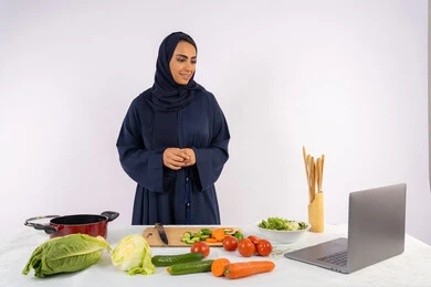 Applying and learning the art of cooking through websites, raising community awareness of preparing healthy food, a portrait of a veiled Emirati Gulf Arab woman wearing an abaya filming and displaying cooking via laptop, a Saudi housewife explaining the ingredients and quantities required via a live broadcast, white background