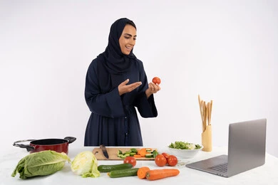 توعية المجتمع بإعداد الطعام الصحي، تعلم وتطبيق فنون الطهي من خلال المواقع الإلكترونية، بورتريه لامرأة عربية خليجية اماراتية محجبة ترتدي العباءة تقوم بتصوير وعرض الطبخ عبر اللابتوب، ربة منزل سعودية تشرح المكونات والكميات المطلوبة عبر بث مباشر، خلفية بيضاء