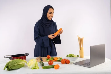 تعلم وتطبيق فنون الطهي من خلال المواقع الإلكترونية، توعية المجتمع بإعداد الطعام الصحي، بورتريه لامرأة عربية خليجية اماراتية محجبة ترتدي العباءة تقوم بتصوير وعرض الطبخ عبر اللابتوب، ربة منزل سعودية تشرح المكونات والكميات المطلوبة عبر بث مباشر، خلفية بيضاء