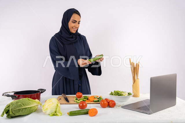 Raising community awareness about preparing healthy food, learning and applying culinary arts through websites, portrait of a veiled Emirati Gulf Arab woman wearing an abaya filming and displaying cooking on a laptop, a Saudi housewife explaining the ingredients and quantities required via a live broadcast, white background
