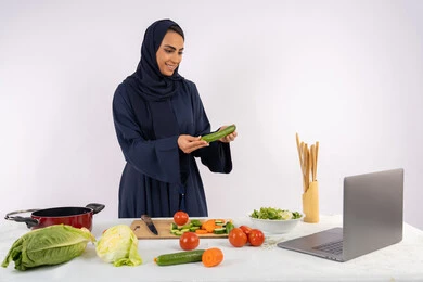 Raising community awareness about preparing healthy food, learning and applying culinary arts through websites, portrait of a veiled Emirati Gulf Arab woman wearing an abaya filming and displaying cooking on a laptop, a Saudi housewife explaining the ingredients and quantities required via a live broadcast, white background