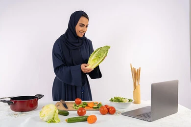 Learning and applying culinary arts through websites, raising community awareness about preparing healthy food, portrait of a veiled Emirati Gulf Arab woman wearing an abaya filming and displaying cooking via laptop, Saudi housewife explaining the ingredients and quantities required via live broadcast, white background