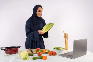 نشر الوعي في المجتمع بإعداد الطعام الصحي، تعلم وتطبيق فنون الطهي من خلال المواقع الإلكترونية، بورتريه لامرأة عربية خليجية اماراتية محجبة ترتدي العباءة تقوم بتصوير وعرض الطبخ عبر اللابتوب، ربة منزل سعودية تشرح المكونات والكميات المطلوبة عبر بث مباشر، خلفية بيضاء