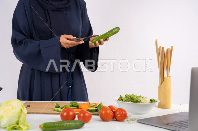 تصوير وصفات الطعام عبر الحاسب المحمول، ربة منزل سعودية تقوم بتحضير وصفة بالخضراوات الطازجة، احترافية اختيار الخضار، بورتريه مقرب لسيدة عربية خليجية اماراتية محجبة ترتدي عباءة تحضر وجبة صحية مفيدة، مشاركة الوصفات على مواقع الانترنت، خلفية بيضاء