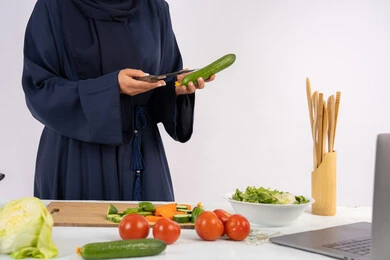 تصوير وصفات الطعام عبر الحاسب المحمول، ربة منزل سعودية تقوم بتحضير وصفة بالخضراوات الطازجة، احترافية اختيار الخضار، بورتريه مقرب لسيدة عربية خليجية اماراتية محجبة ترتدي عباءة تحضر وجبة صحية مفيدة، مشاركة الوصفات على مواقع الانترنت، خلفية بيضاء