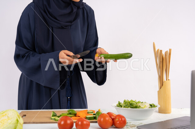 مشاركة الوصفات على مواقع الانترنت، تعلم مهارة الطهي وإعداد الطعام عبر الحاسب المحمول، ربة منزل سعودية تقوم بتحضير وصفة بالخضراوات الطازجة، احترافية اختيار الخضار، بورتريه مقرب لسيدة عربية خليجية اماراتية محجبة ترتدي عباءة تحضر وجبة صحية مفيدة، خلفية بيضاء