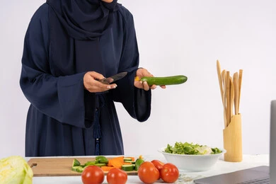 مشاركة الوصفات على مواقع الانترنت، تعلم مهارة الطهي وإعداد الطعام عبر الحاسب المحمول، ربة منزل سعودية تقوم بتحضير وصفة بالخضراوات الطازجة، احترافية اختيار الخضار، بورتريه مقرب لسيدة عربية خليجية اماراتية محجبة ترتدي عباءة تحضر وجبة صحية مفيدة، خلفية بيضاء
