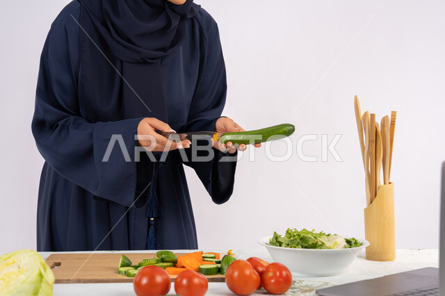 تعلم مهارة الطهي وإعداد الطعام عبر الحاسب المحمول، ربة منزل سعودية تقوم بتحضير وصفة بالخضراوات الطازجة، احترافية اختيار الخضار، بورتريه مقرب لسيدة عربية خليجية اماراتية محجبة ترتدي عباءة تحضر وجبة صحية مفيدة، مشاركة الوصفات على مواقع الانترنت، خلفية بيضاء