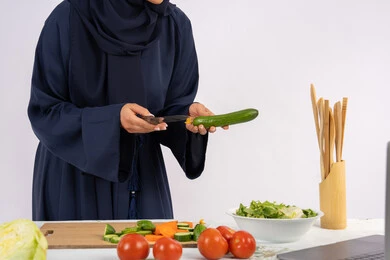 تعلم مهارة الطهي وإعداد الطعام عبر الحاسب المحمول، ربة منزل سعودية تقوم بتحضير وصفة بالخضراوات الطازجة، احترافية اختيار الخضار، بورتريه مقرب لسيدة عربية خليجية اماراتية محجبة ترتدي عباءة تحضر وجبة صحية مفيدة، مشاركة الوصفات على مواقع الانترنت، خلفية بيضاء