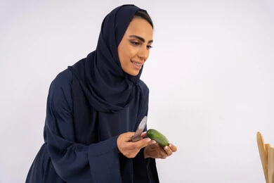وجبة صحية مفيدة، تعلم مهارة الطهي وإعداد الطعام، ربة منزل سعودية تقوم بتحضير وصفة بالخضراوات الطازجة، احترافية اختيار الخضار، بورتريه مقرب لسيدة عربية خليجية اماراتية محجبة ترتدي عباءة تمسك بيدها الخيار، خلفية بيضاء