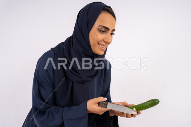 وجبة صحية مفيدة، تعلم مهارة الطهي وإعداد الطعام، ربة منزل سعودية تقوم بتحضير وصفة بالخضراوات الطازجة، احترافية اختيار الخضار، بورتريه مقرب لسيدة عربية خليجية اماراتية محجبة ترتدي عباءة تمسك بيدها الخيار، خلفية بيضاء