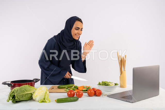 Raising hands with greeting gestures, learning and practicing cooking arts through websites, portrait of a veiled Emirati Gulf Arab woman wearing an abaya filming and displaying cooking on a laptop, a Saudi housewife explaining the ingredients and quantities required via a live broadcast, white background