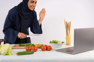 The importance of healthy food, greeting and saluting, learning and practicing the art of cooking through websites, a portrait of a veiled Emirati Gulf Arab woman wearing an abaya filming and displaying cooking via laptop, a Saudi housewife explaining the ingredients and quantities required via a live broadcast, white background