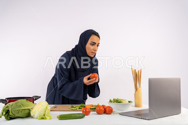 Raising community awareness about preparing healthy food, learning and applying culinary arts through websites, portrait of a veiled Emirati Gulf Arab woman wearing an abaya filming and displaying cooking on a laptop, a Saudi housewife explaining the ingredients and quantities required via a live broadcast, white background