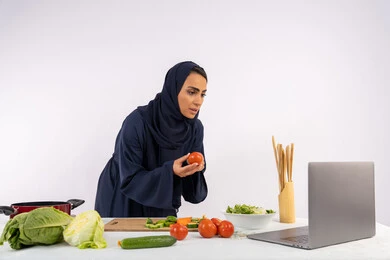 Raising community awareness about preparing healthy food, learning and applying culinary arts through websites, portrait of a veiled Emirati Gulf Arab woman wearing an abaya filming and displaying cooking on a laptop, a Saudi housewife explaining the ingredients and quantities required via a live broadcast, white background