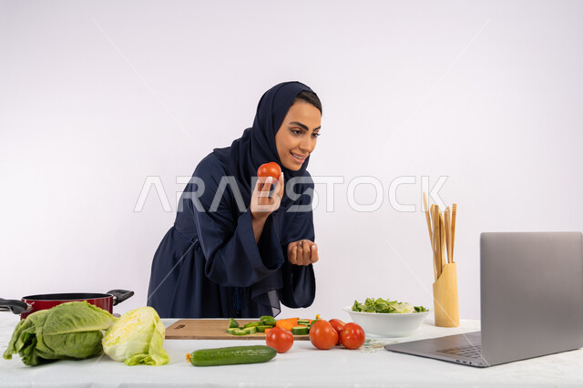 تعلم وتطبيق فنون الطهي من خلال المواقع الإلكترونية، توعية المجتمع بإعداد الطعام الصحي، بورتريه لامرأة عربية خليجية اماراتية محجبة ترتدي العباءة تقوم بتصوير وعرض الطبخ عبر اللابتوب، ربة منزل سعودية تشرح المكونات والكميات المطلوبة عبر بث مباشر، خلفية بيضاء