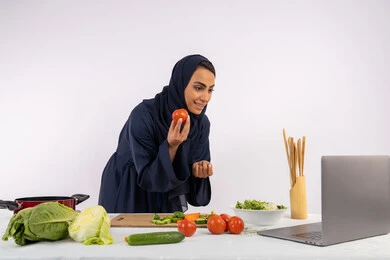 Learning and applying culinary arts through websites, raising community awareness about preparing healthy food, portrait of a veiled Emirati Gulf Arab woman wearing an abaya filming and displaying cooking via laptop, Saudi housewife explaining the ingredients and quantities required via live broadcast, white background
