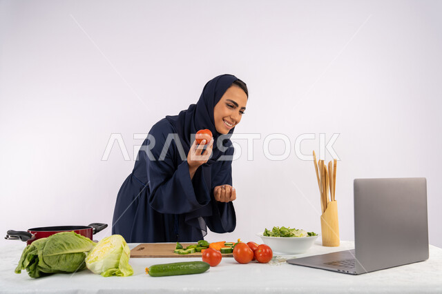 توعية المجتمع بإعداد الطعام الصحي، تعلم وتطبيق فنون الطهي من خلال المواقع الإلكترونية، بورتريه لامرأة عربية خليجية اماراتية محجبة ترتدي العباءة تقوم بتصوير وعرض الطبخ عبر اللابتوب، ربة منزل سعودية تشرح المكونات والكميات المطلوبة عبر بث مباشر، خلفية بيضاء