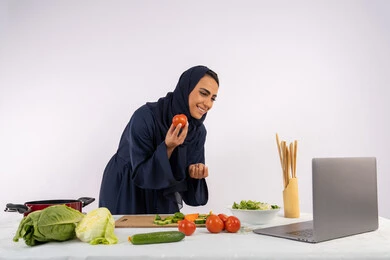 توعية المجتمع بإعداد الطعام الصحي، تعلم وتطبيق فنون الطهي من خلال المواقع الإلكترونية، بورتريه لامرأة عربية خليجية اماراتية محجبة ترتدي العباءة تقوم بتصوير وعرض الطبخ عبر اللابتوب، ربة منزل سعودية تشرح المكونات والكميات المطلوبة عبر بث مباشر، خلفية بيضاء