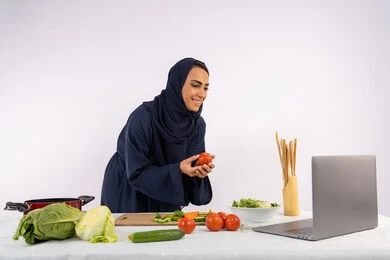 تعلم وتطبيق فنون الطهي من خلال المواقع الإلكترونية، توعية المجتمع بإعداد الطعام الصحي، بورتريه لامرأة عربية خليجية اماراتية محجبة ترتدي العباءة تقوم بتصوير وعرض الطبخ عبر اللابتوب، ربة منزل سعودية تشرح المكونات والكميات المطلوبة عبر بث مباشر، خلفية بيضاء