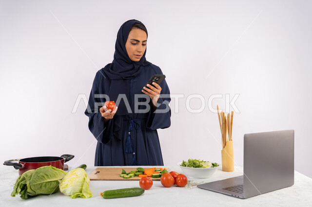 إعداد السلطة الصحية من الخضراوات الطازجة، تعلم مهارة الطهي عبر الحاسوب، بورتريه لسيدة عربية خليجية اماراتية محجبة  ترتدي عباءة  تقوم بتحضير وصفة طبق صحي مفيد من الانترنت، ربة منزل سعودية تستخدم الهاتف المحمول، خلفية بيضاء