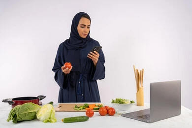 Preparing a healthy salad from fresh vegetables, learning cooking skills via computer, portrait of a veiled Emirati Gulf Arab woman wearing an abaya preparing a healthy and useful dish recipe from the internet, a Saudi housewife using a mobile phone, white background