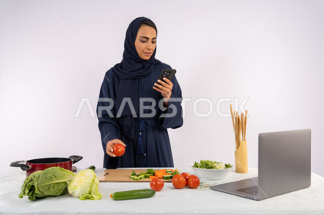 تعابير الاندماج والتركيز في الموبايل، إعداد السلطة الصحية من الخضراوات الطازجة، تعلم مهارة الطهي عبر الحاسوب، بورتريه لسيدة عربية خليجية اماراتية محجبة  ترتدي عباءة  تقوم بتحضير وصفة طبق صحي مفيد من الانترنت، ربة منزل سعودية تستخدم الهاتف المحمول، خلفية بيضاء