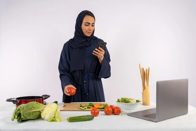 Expressions of immersion and concentration on the mobile phone, preparing a healthy salad with fresh vegetables, learning cooking skills via computer, portrait of a veiled Emirati Gulf Arab woman wearing an abaya preparing a healthy and useful dish recipe from the internet, a Saudi housewife using a mobile phone, white background