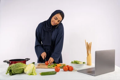 Talking and making a call on a mobile phone, preparing a healthy salad, learning cooking skills via computer, portrait of a veiled Emirati Gulf Arab woman wearing an abaya preparing a healthy dish recipe from the internet, a Saudi housewife cutting fresh vegetables, white background