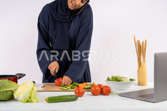 إجراء مكالمة عبر الهاتف الجوال، إعداد السلطة الصحية، تعلم مهارة الطهي عبر الحاسوب، بورتريه لسيدة عربية خليجية اماراتية محجبة  ترتدي عباءة تقوم بتحضير وصفة طبق صحي مفيد من الانترنت، ربة منزل سعودية تقوم بتقطيع الخضراوات الطازجة، خلفية بيضاء