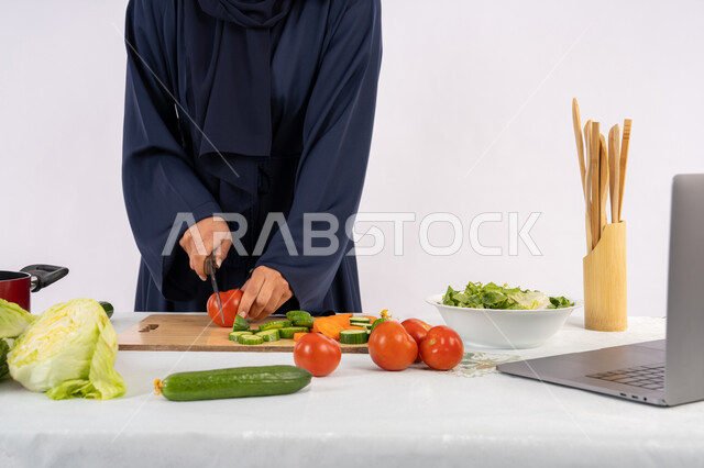 Learning the skill of cooking and preparing food via laptop, portrait of a veiled Emirati Gulf Arab woman wearing an abaya preparing a recipe for a healthy and useful dish from the internet, a Saudi housewife cutting fresh vegetables, using the appropriate ingredients and quantities for the meal, white background