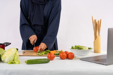تعلم مهارة الطهي وإعداد الطعام عبر الحاسب المحمول، بورتريه لسيدة عربية خليجية اماراتية محجبة  ترتدي عباءة تقوم بتحضير وصفة طبق صحي مفيد من الانترنت، ربة منزل سعودية تقوم بتقطيع الخضراوات الطازجة، استخدام المكونات والمقادير المناسبة للوجبة، خلفية بيضاء