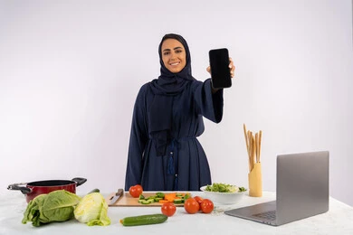 ربة منزل سعودية تعرض شاشة سوداء فارغة عبر الهاتف المحمول بتعابير السرور، إعداد السلطة الصحية من الخضراوات الطازجة، تعلم مهارة الطهي عبر الحاسوب، بورتريه لسيدة عربية خليجية اماراتية محجبة  ترتدي عباءة  تقوم بتحضير وصفة طبق صحي مفيد من الانترنت، خلفية بيضاء