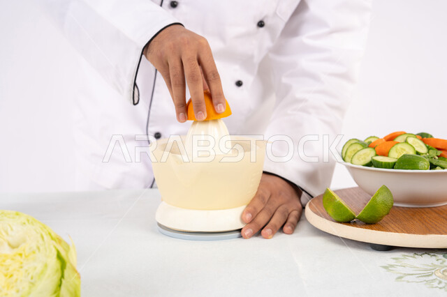 مفهوم النظام الغذائي الصحي، شيف سعودي يقوم بتحضير عصير مفيد، شغف تعلم مهارة الطهي، اعداد المشروبات الصحية والطازجة، بورتريه مقرب لرجل عربي خليجي اماراتي يرتدي زي الطهاة يعصر البرتقال الطازج، خلفية بيضاء