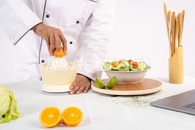 طباخ سعودي يقوم بتحضير عصير مفيد، شغف تعلم مهارة الطهي، مفهوم النظام الغذائي الصحي، اعداد المشروبات الصحية والطازجة، بورتريه مقرب لرجل عربي خليجي اماراتي يرتدي زي الطهاة يعصر البرتقال الطازج، خلفية بيضاء