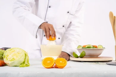 طاهي سعودي يقوم بتحضير عصير مفيد، شغف تعلم مهارة الطهي، مفهوم النظام الغذائي الصحي، اعداد المشروبات الصحية والطازجة، بورتريه مقرب لرجل عربي خليجي اماراتي يرتدي زي الطهاة يعصر البرتقال الطازج، خلفية بيضاء