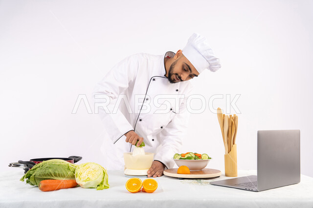 Publish and share recipes on websites, a Saudi chef preparing a healthy juice, learning cooking skills via laptop, the concept of a healthy diet, preparing healthy and fresh drinks, a portrait of an Arab Gulf Emirati man wearing a chef's uniform squeezing fresh oranges, white background