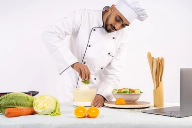 تصوير ومشاركة الوصفات على مواقع الانترنت، شيف سعودي يقوم بتحضير عصير مفيد، تعلم مهارة الطهي عبر الحاسوب المحمول، مفهوم النظام الغذائي الصحي، اعداد المشروبات الصحية والطازجة، بورتريه لرجل عربي خليجي اماراتي يرتدي زي الطهاة يعصر البرتقال الطازج، خلفية بيضاء