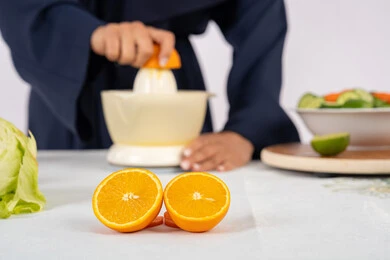 ربة منزل سعودية تقوم بتحضير عصير مفيد، مفهوم النظام الغذائي الصحي، حب تعلم مهارة الطهي، اعداد المشروبات الصحية والطازجة، بورتريه مقرب لسيدة عربية خليجية اماراتية ترتدي عباءة تعصر البرتقال الطازج، خلفية بيضاء