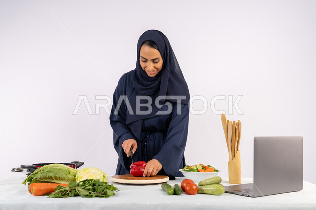 Preparing salads and healthy dishes, learning cooking skills via laptop, portrait of a veiled Emirati Gulf Arab woman wearing an abaya preparing a healthy dish recipe from the internet, a Saudi housewife cutting fresh vegetables, white background
