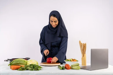 Preparing salads and healthy dishes, learning cooking skills via laptop, portrait of a veiled Emirati Gulf Arab woman wearing an abaya preparing a healthy dish recipe from the internet, a Saudi housewife cutting fresh vegetables, white background