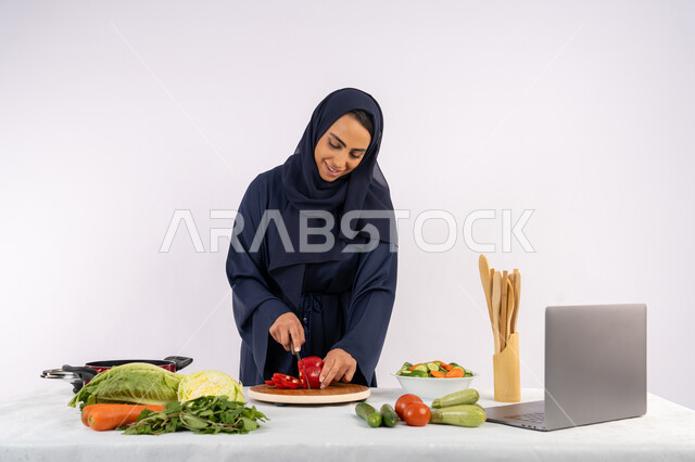 اتباع نظام غذائي صحي، إعداد السلطات والاطباق الصحية، تعلم مهارة الطهي عبر الحاسب المحمول، بورتريه لسيدة عربية خليجية اماراتية محجبة  ترتدي عباءة تقوم بتحضير وصفة طبق مفيد من الانترنت، ربة منزل سعودية تقوم بتقطيع الخضراوات الطازجة، خلفية بيضاء