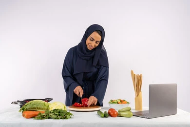 اتباع نظام غذائي صحي، إعداد السلطات والاطباق الصحية، تعلم مهارة الطهي عبر الحاسب المحمول، بورتريه لسيدة عربية خليجية اماراتية محجبة  ترتدي عباءة تقوم بتحضير وصفة طبق مفيد من الانترنت، ربة منزل سعودية تقوم بتقطيع الخضراوات الطازجة، خلفية بيضاء