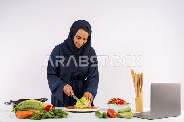 salads and healthy dishes, learning cooking skills via laptop, portrait of a veiled Emirati Gulf Arab woman wearing an abaya preparing a healthy dish recipe from the internet, a Saudi housewife cutting fresh vegetables, white background