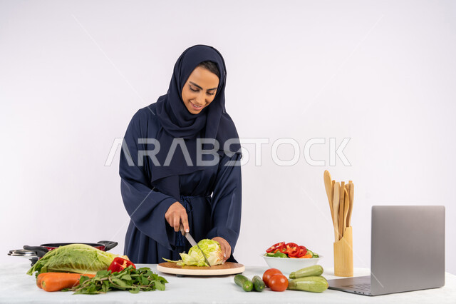 إعداد السلطات والاطباق الصحية، تعلم مهارة الطهي عبر الحاسب المحمول، بورتريه لسيدة عربية خليجية اماراتية محجبة  ترتدي عباءة تقوم بتحضير وصفة طبق صحي مفيد من الانترنت، ربة منزل سعودية تقوم بتقطيع الخضراوات الطازجة، خلفية بيضاء