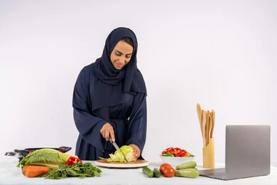 Preparing salads and healthy dishes, learning cooking skills via laptop, portrait of a veiled Emirati Gulf Arab woman wearing an abaya preparing a healthy dish recipe from the internet, a Saudi housewife cutting fresh vegetables, white background