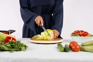 اتباع نظام غذائي صحي، إعداد السلطات والاطباق الصحية، شغف تعلم مهارة الطهي، بورتريه مقرب لسيدة عربية خليجية اماراتية ترتدي عباءة تقوم بتحضير طبق مفيد، ربة منزل سعودية تقوم بتقطيع الخضراوات الطازجة، خلفية بيضاء